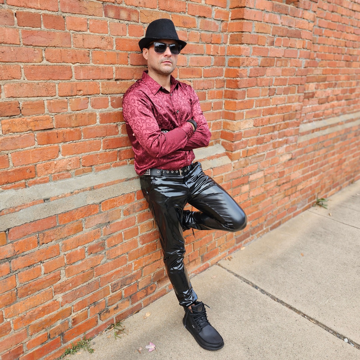 Cosmicus in city standing on brick wall in shiny faux leather pvc pants and boots